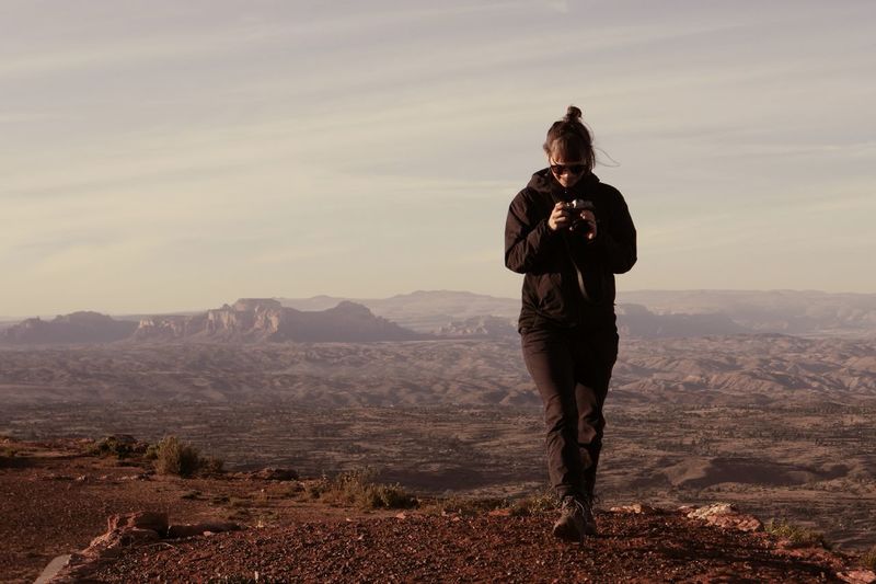 Full length of woman holding camera while walking on mountain against sky