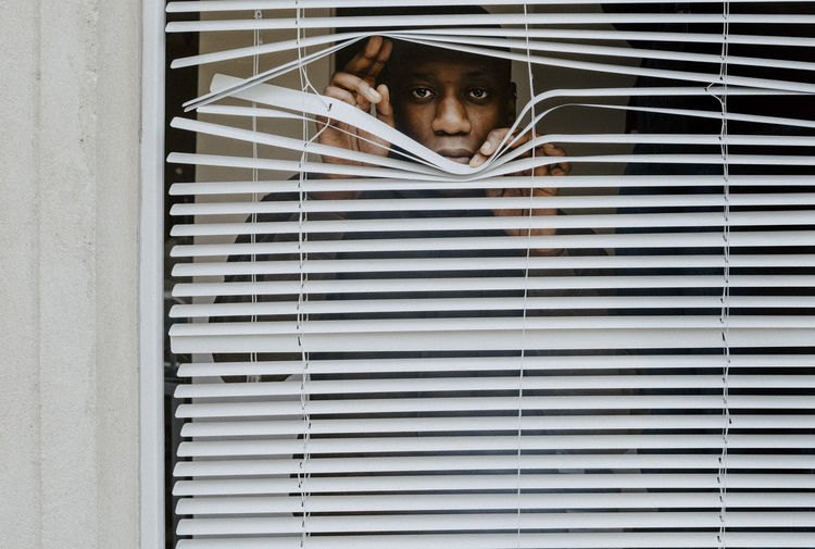 Young man looking out of window blinds at home