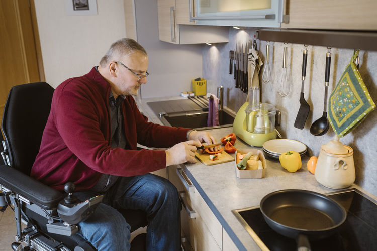 Retired senior man with disability sitting in motorized wheelchair cutting bell pepper at kitchen counter