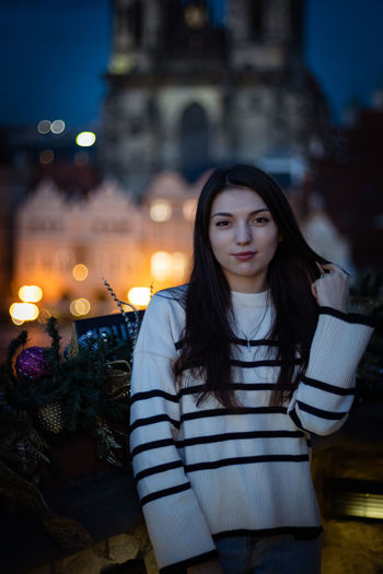 Portrait of young woman standing against buildings in city at night