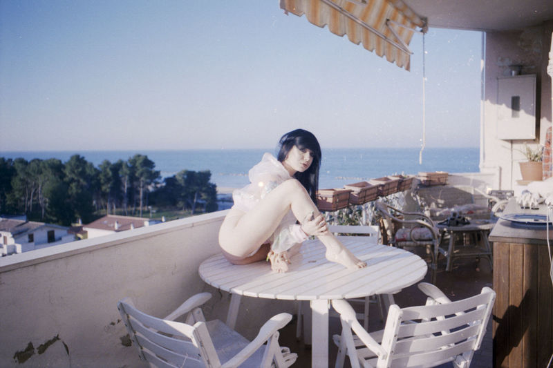Young woman sitting on chair by sea against clear sky