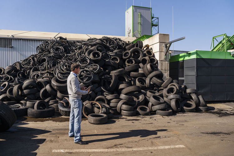 Mature businessman with tablet pc standing by heap of rubber tires at recycling plant