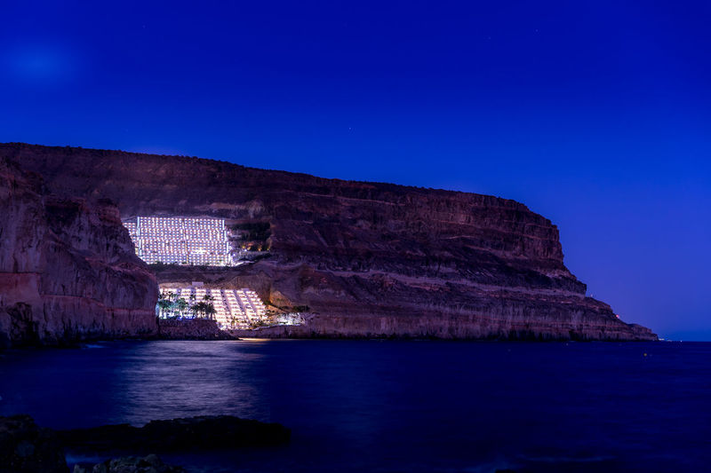 View of illuminated buildings on rock formation by sea against sky