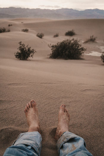 Male feet on soft desert sand sitting on a dune in desert at sunset with mountains and dramatic sky.