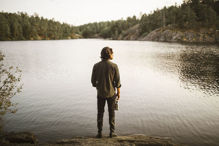 Rear view of man with bottle standing by lake in forest during vacation