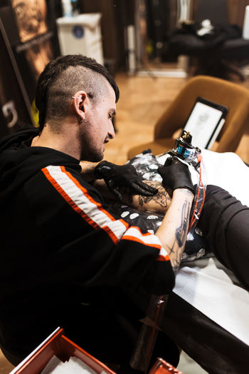 From above stylish man with piercing using tattoo machine to make tattoo on leg of crop customer during work in salon
