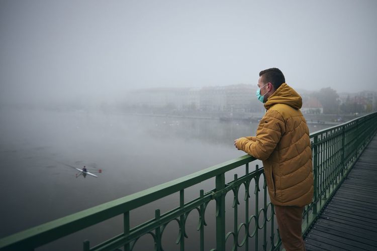 Man wearing mask standing by railing on bridge against sky during winter