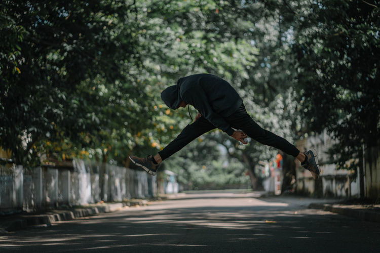 Side view of young man jumping on road against trees