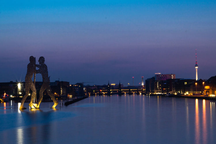Molecule men sculpture on river in city against sky at night