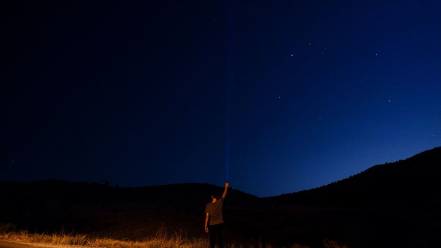 Rear view of man holding illuminated flashlight against sky at night