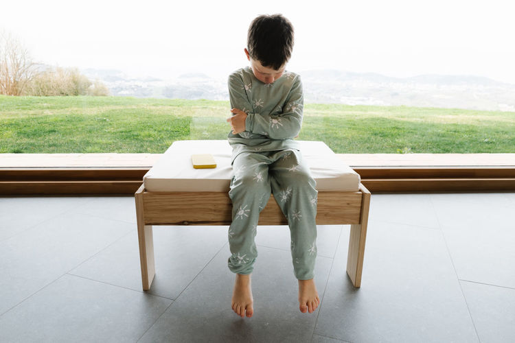 Unhappy boy in pajama sitting on pouf in light room on morning time near modern cellphone to faceless