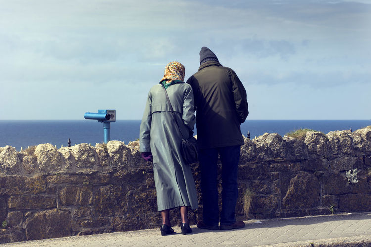 Rear view of couple looking at sea while standing by retaining wall against sky