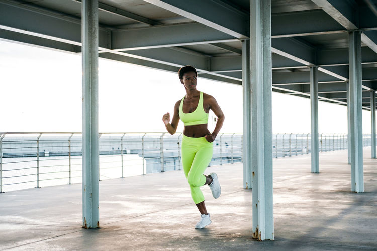 From below african american adult sportswoman in vibrant green activewear focusing and running alone along waterfront among metal columns under roof
