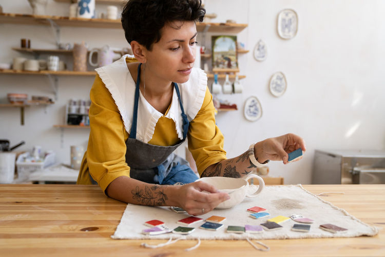 Young female ceramist in apron working in pottery studio, choosing paint color for ceramics goods