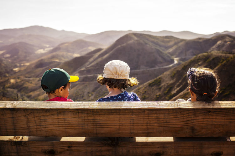 Rear view of siblings sitting on bench against mountains