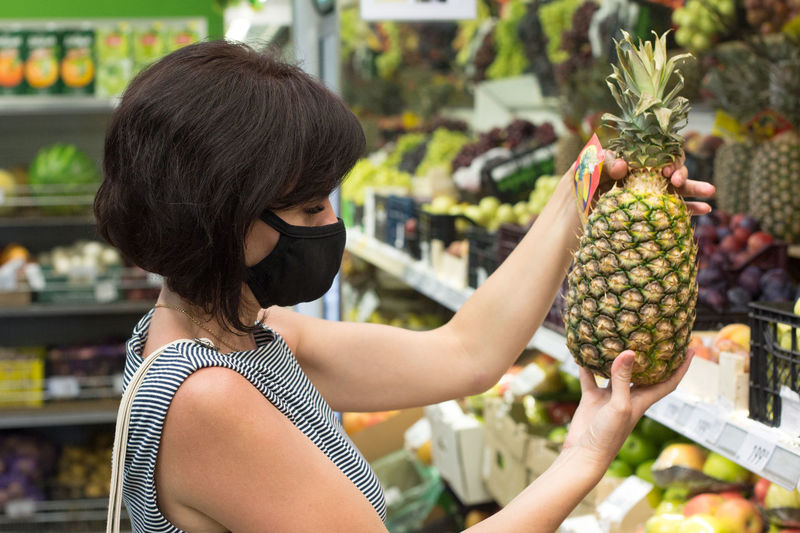 Woman buyer protective medical mask chooses a pineapple in a store. person