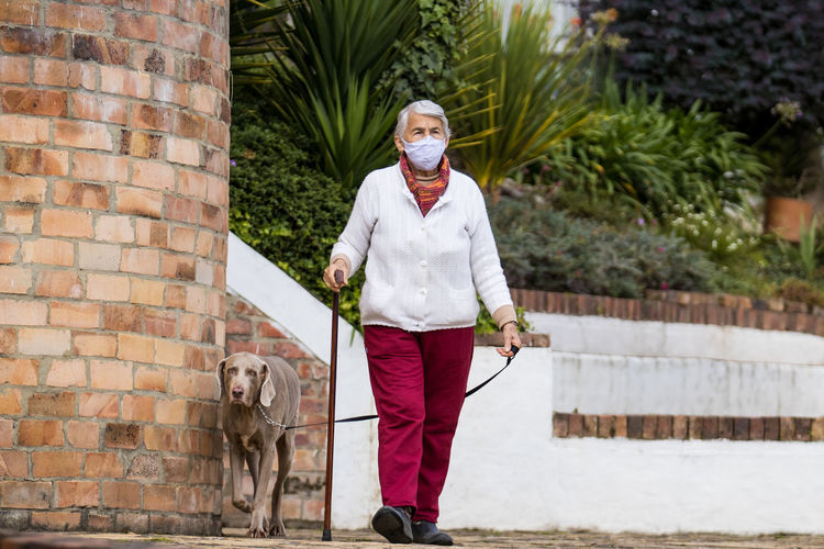 Full length of senior wearing mask with dog walking outdoors