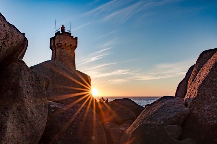 Lighthouse amidst rocks and sea against sky during sunset