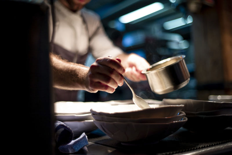 Hands of cook holding spoon and little pan while adding sauce in plate on restaurant kitchen
