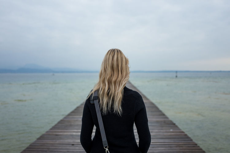 Rear view of mid adult woman standing on pier over sea against cloudy sky