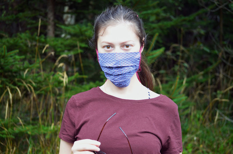 Portrait of young woman with brown ponytail in a colorful patterned face mask on a nature background