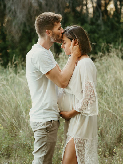 Side view of tender male kissing pregnant female in forehead while standing in field in nature