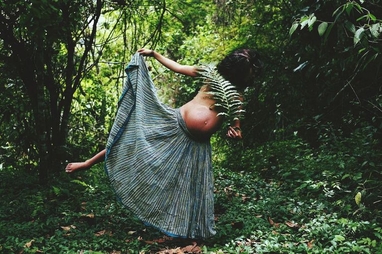 Shirtless pregnant woman holding leaves while standing in forest