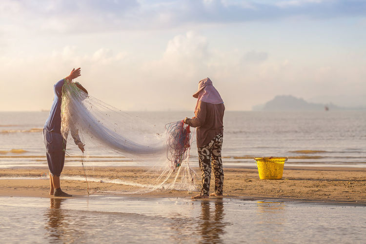 Man and woman holding fishing net standing on beach against sky