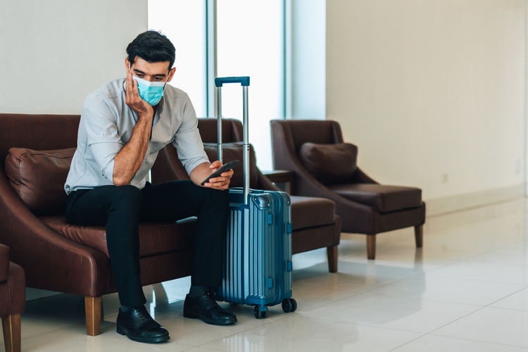 Young businessman wearing mask with suitcase sitting at airport lounge