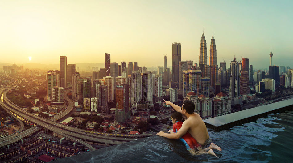 Shirtless father with daughter in swimming pool pointing at cityscape during sunset