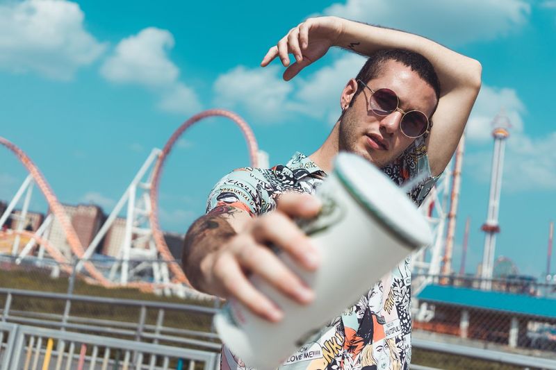 Portrait of young man wearing sunglasses holding coffee while standing against amusement park
