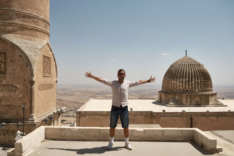 Young adult man stands posing with opened arms on a roof against ulu cami or grand mosque of mardin