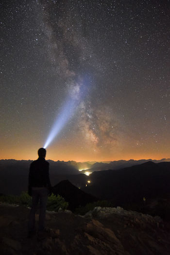 Man pointing flashlight towards star field while standing on mountain peak at night