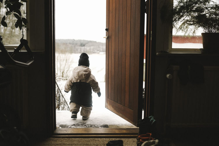 Full length rear view of baby girl in warm clothing walking out from doorway during winter