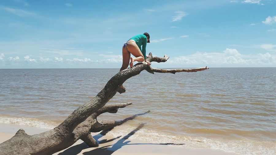 Full length of woman on driftwood at beach against sky