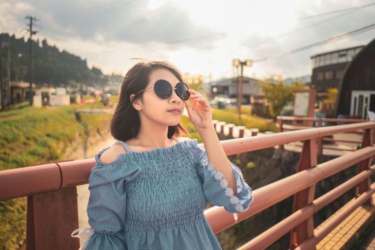Young woman wearing sunglasses standing on bridge against railing