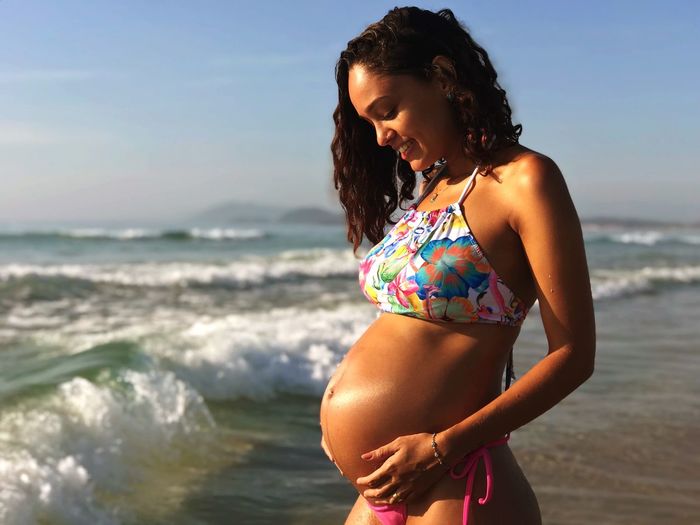 Pregnant woman wearing bikini while standing at beach against blue sky