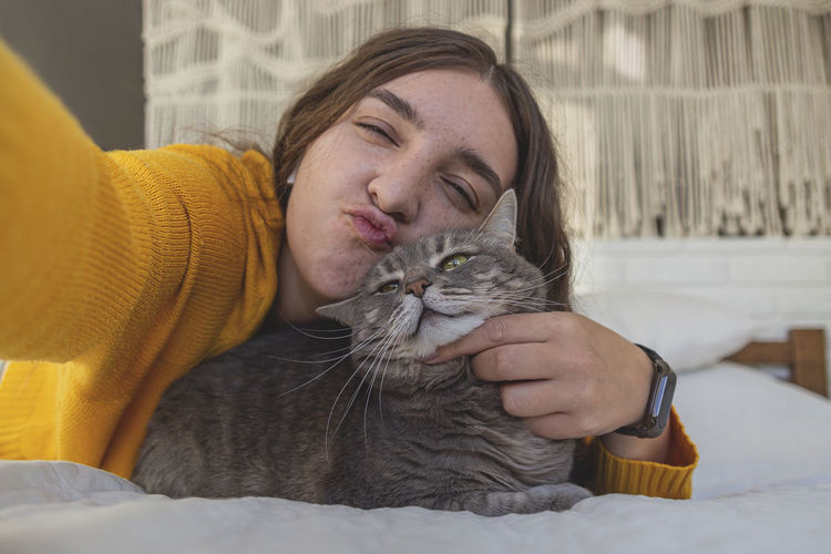 Happy woman in yellow sweater hugs her gray cat on the bed in a light interior