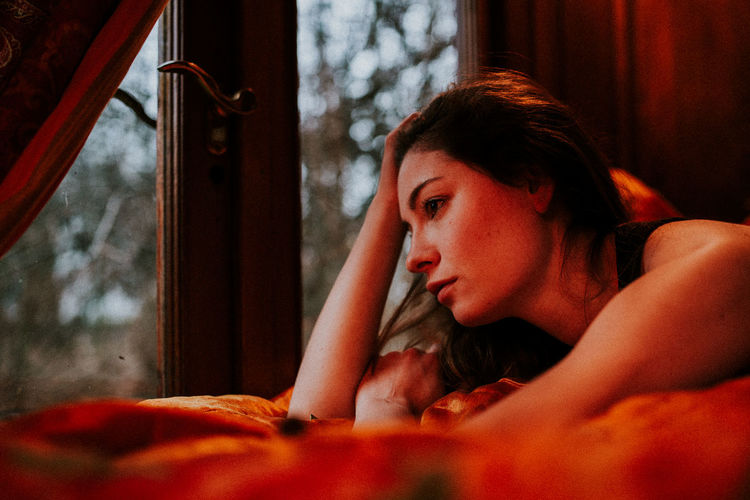 Close-up of thoughtful woman with hand in hair lying on bed by window at home