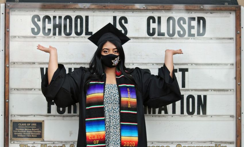 Portrait of young woman wearing mask and graduation gown standing against wall