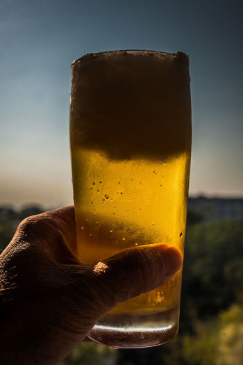 Close-up of hand holding beer glass against sunset