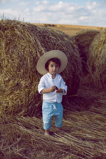 Boy a child in a straw hat and blue pants stands in a mowed field with stacks in the summer