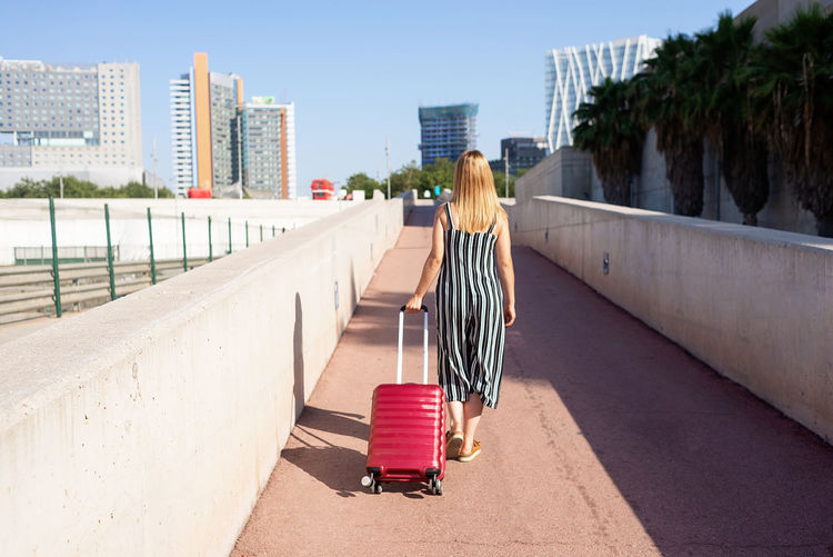 Rear view of a young traveler woman carrying a red trolley in the city