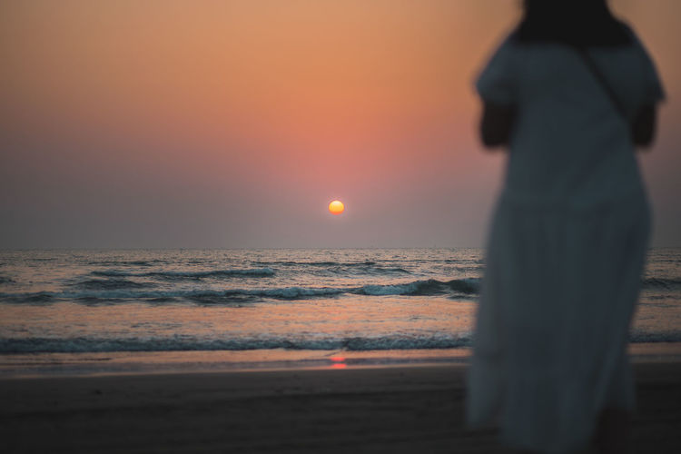 Rear view of woman standing at beach during sunset