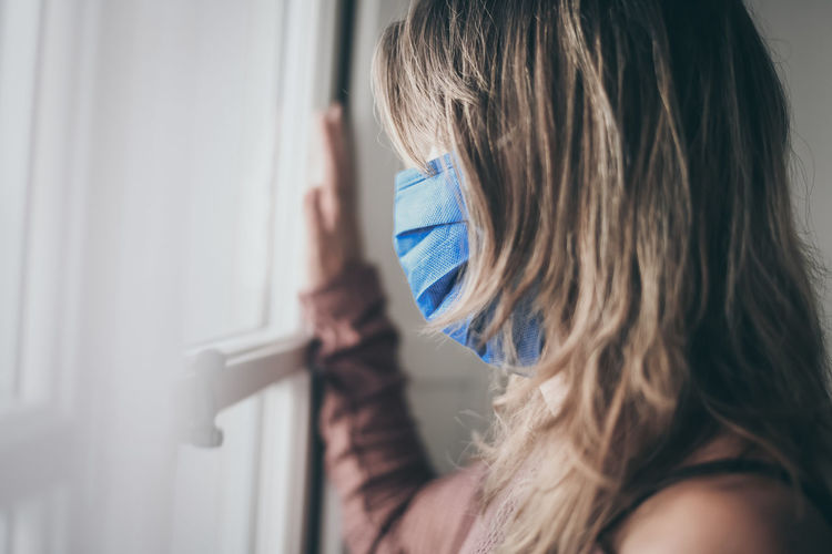 Young woman looking outside window with surgical mask during coronavirus lockdown covid-19 isolated
