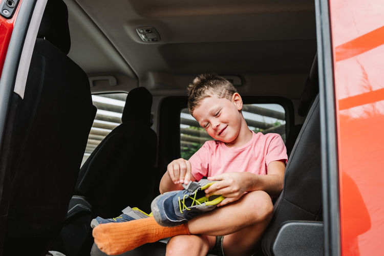 Cheerful boy in casual clothes fastening shoe while sitting on back seat of red vehicle during road trip