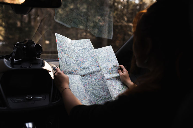 Unrecognizable female traveler sitting on passenger seat and reading tourist map while checking route during road trip through woodland