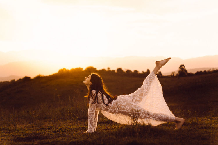 Side view of young woman dancing on grassy field against sky during sunset