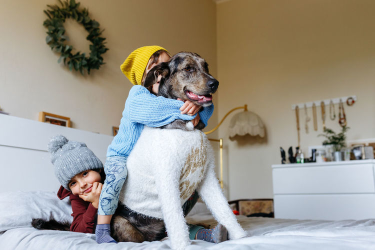 Two children wearing warm hats hug their dog which is wearing sweater