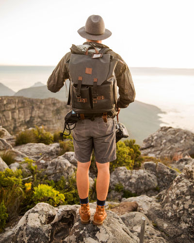 Full length rear view of man with backpack walking on rocky mountains against sea